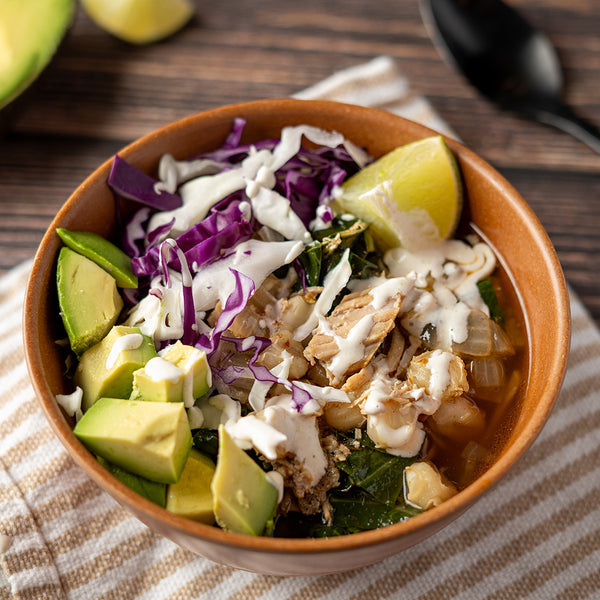 A bowl of braised pork and hominy stew garnished with avocado, lime, red cabbage, and a drizzle of sauce, on a patterned napkin.