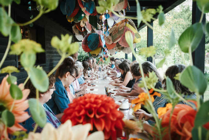 Campers working side by side at a long table on the covered porch at Iola Gardens, with dahlias in the foreground and paper decorations hanging overhead