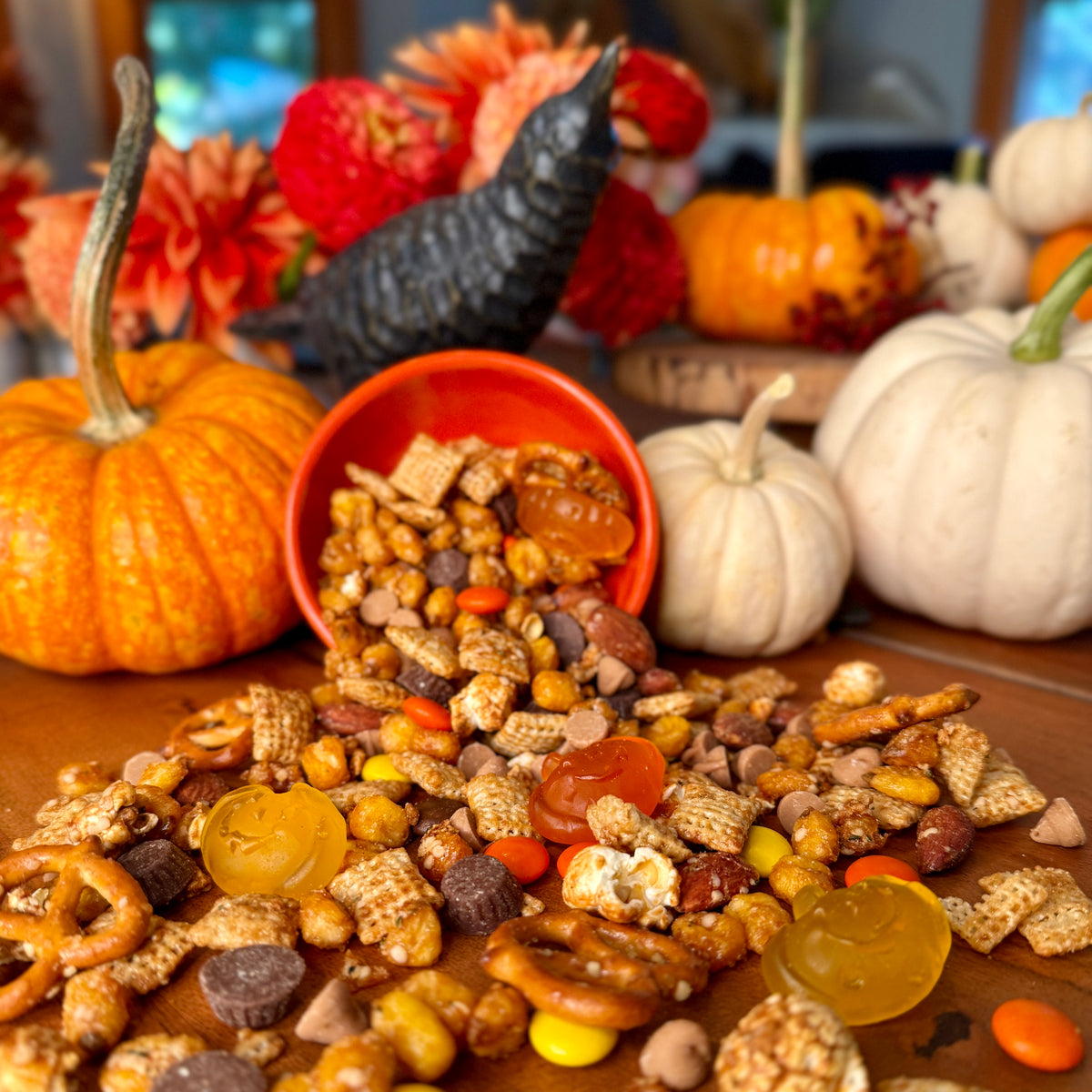 A variety of snacks including Chex, pretzels, corn, almonds, gummy apples, Reese's Pieces, mini peanut butter cups, and sea salt caramel chips arranged on a table with decorative pumpkins and autumn leaves in the background.