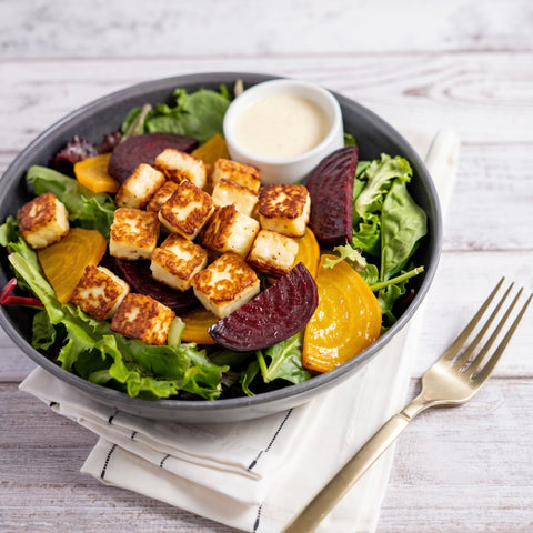 A prepared salad in a dark bowl with grilled cubed halloumi, green lettuce, beet slices, orange beets, and a small white cup of creamy dressing, styled on a white napkin with a gold fork on a light wood surface.