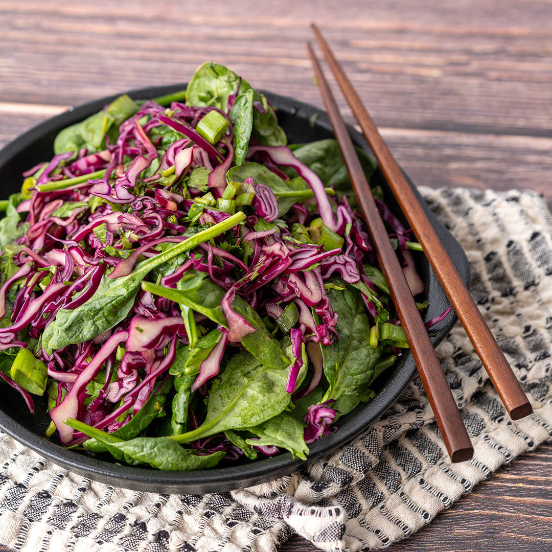A bowl of Asian Sesame Spinach Slaw with red cabbage and green spinach, accompanied by chopsticks, set on a patterned napkin.