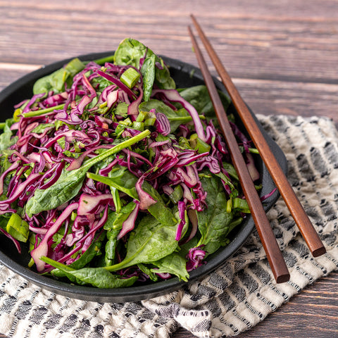 A bowl of Asian Sesame Spinach Slaw with red cabbage and green spinach, accompanied by chopsticks, set on a patterned napkin.
