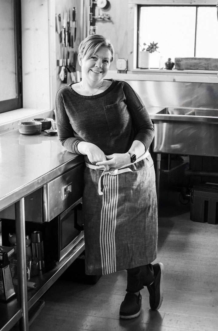 Heidi Finley in an apron, smiling, leaning against a stainless prep bench in the Maven Meals kitchen with a knife rack on the wall behind her