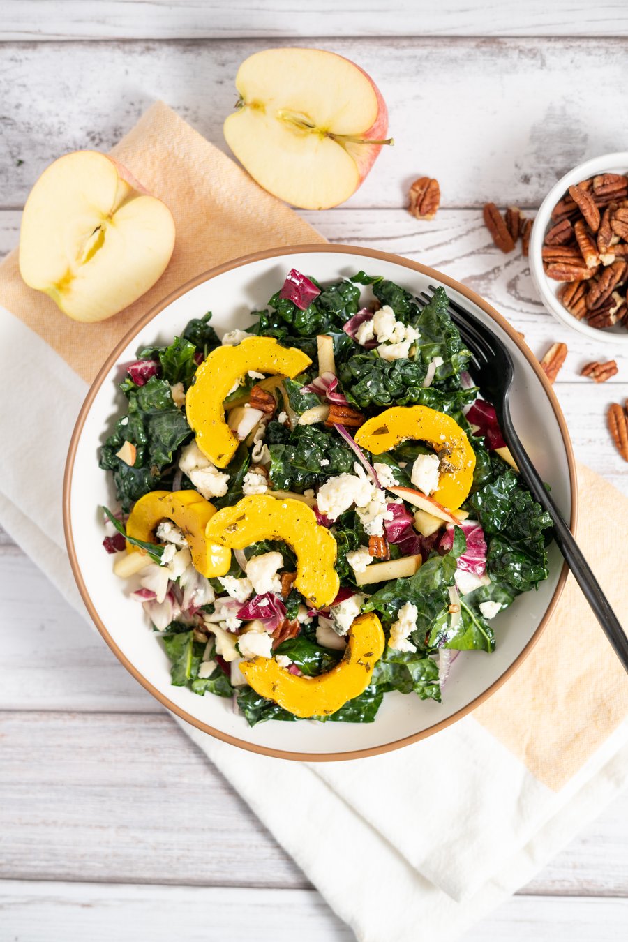 A kale salad with delicata squash, apple, blue cheese and pecans in a white bowl