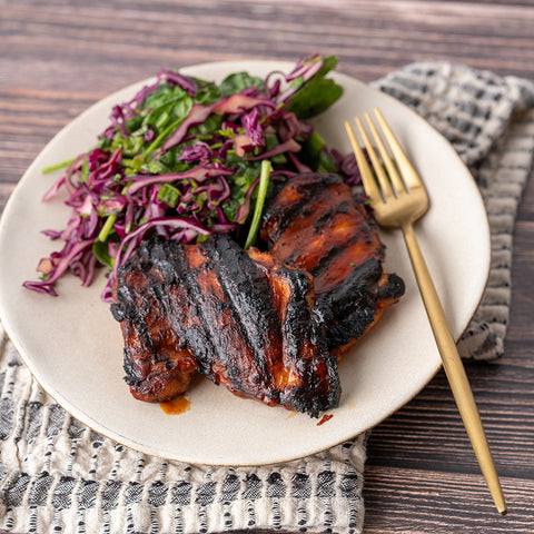 A plate with Korean BBQ chicken thighs and wasabi mashed potatoes, accompanied by a side of leafy greens, served on a wooden table with a striped napkin.