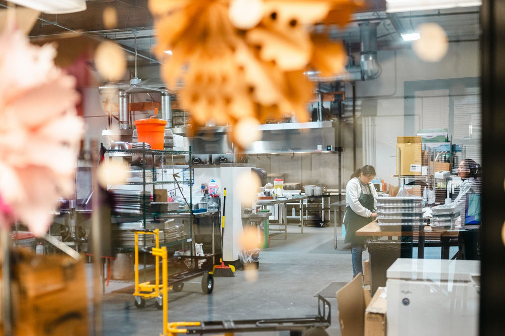 The Maven kitchen near Angle Lake seen through a window, with two cooks packing stacks of foil trays at a wooden table