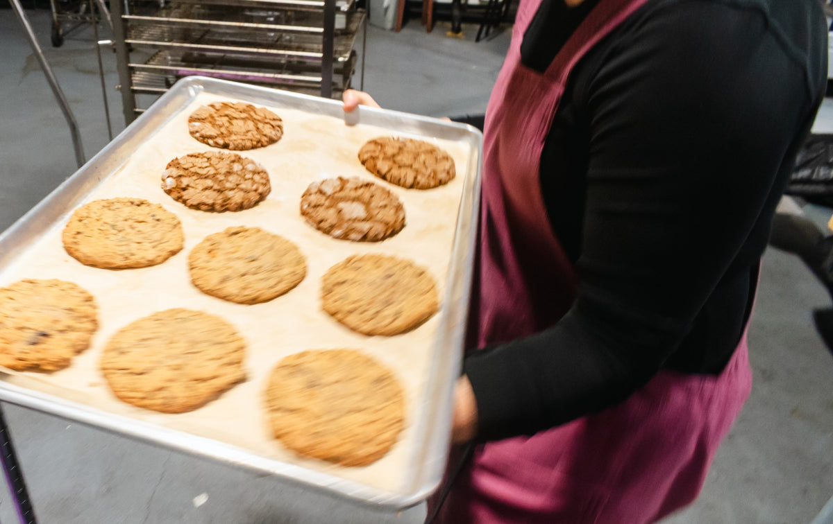 A Maven Meals baker in a burgundy apron carries a sheet tray of fresh-baked oatmeal and crinkle cookies through the SeaTac kitchen