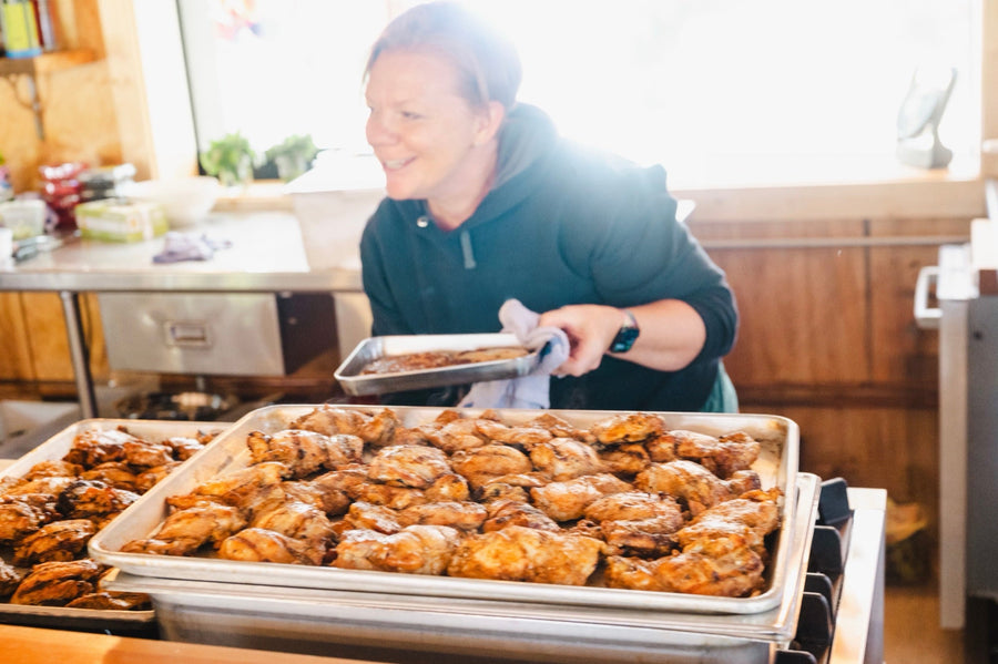 Heidi Finley, founder of Maven Meals, smiles as she carries a pan past full trays of grilled chicken thighs in the kitchen