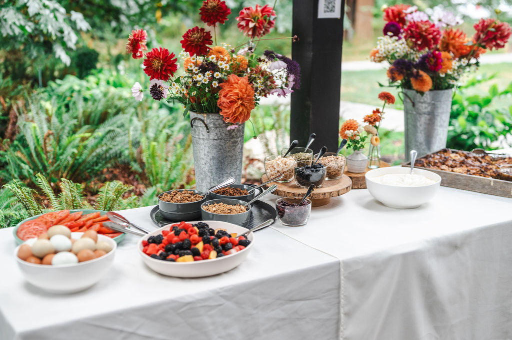 An outdoor Maven Meals catering table laid with a white cloth: granola, yogurt, mixed berries, sliced heirloom tomatoes, a bowl of eggs and a tray of pastries, with dahlias in galvanized buckets behind it