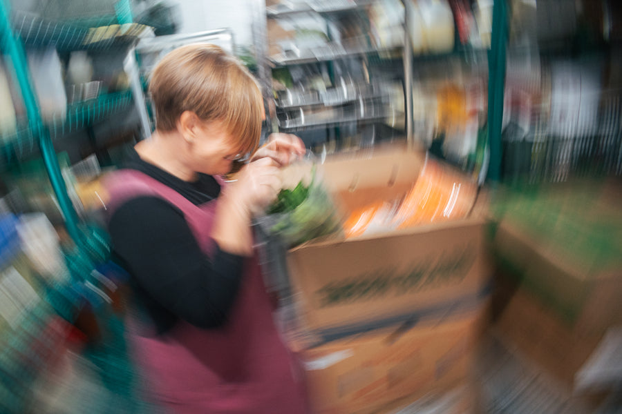 A Maven Meals cook in a burgundy apron leans in over a container of fresh greens beside a box of produce, with the busy SeaTac kitchen blurred in motion all around