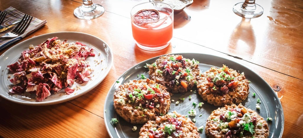 A radicchio salad and a plate of grain cakes topped with a tomato and herb relish, set on a wooden table beside a blood orange drink