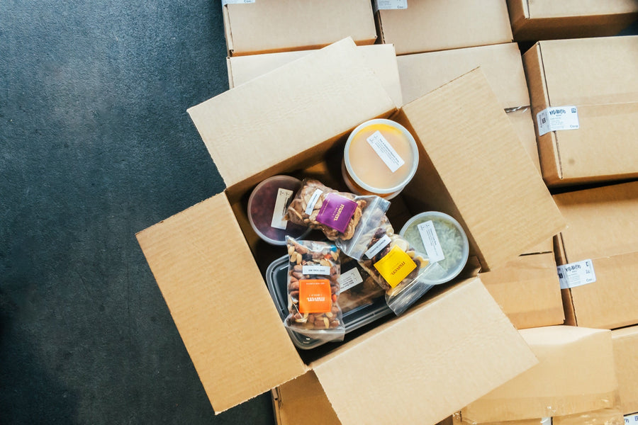 An open Maven Meals delivery box, seen from above, packed with soup, labeled containers and bags of Maven trail mix, beside stacked boxes ready for delivery