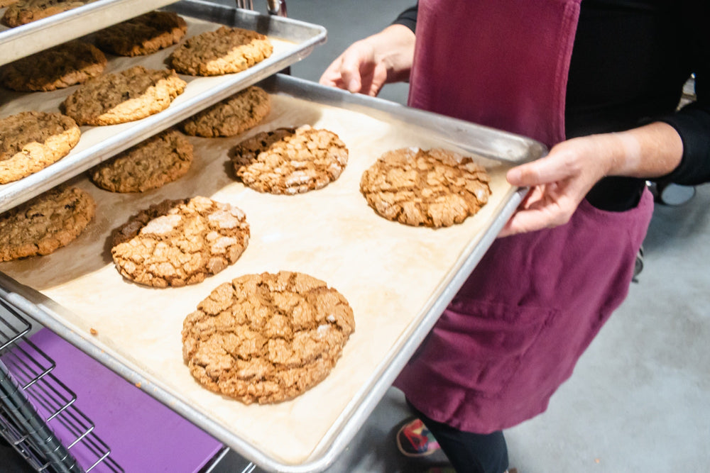 A baker in a burgundy apron slides a tray of crackled molasses cookies onto a rack of fresh-baked cookies
