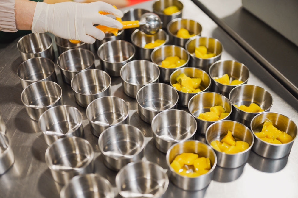 A gloved hand scoops diced yellow fruit into rows of small stainless steel cups for school lunches