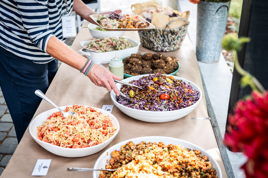 A Maven Meals serve line of salads, grain bowls and slaw, being plated by hand