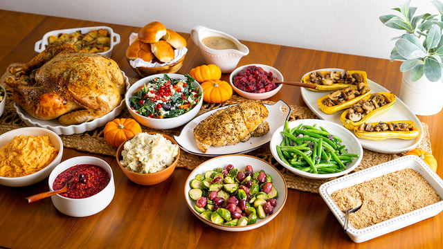 A Thanksgiving table with a whole roast turkey, a sliced herb-roasted turkey breast, mashed potatoes, sweet potatoes, cranberry sauce, green beans, Brussels sprouts with grapes, mushroom-stuffed squash, a kale salad, stuffing, rolls and gravy.