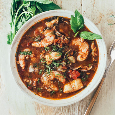 A bowl of seafood and sausage cioppino with a spoon on the side, garnished with herbs.