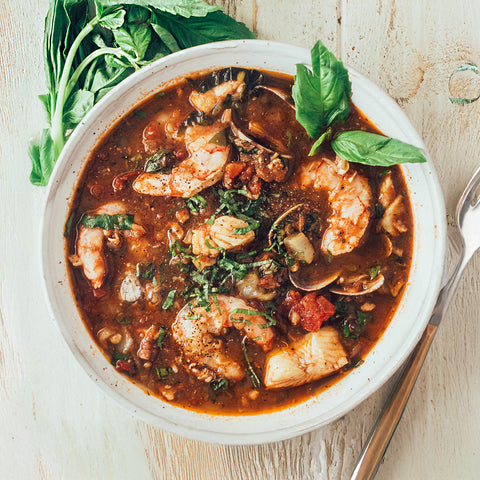 A bowl of seafood and sausage cioppino with a spoon on the side, garnished with herbs.