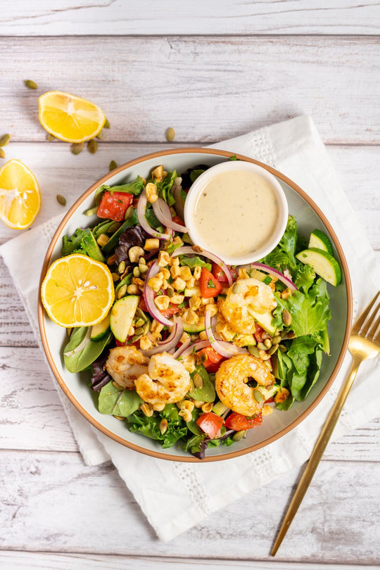 An overhead bowl of shrimp salad with greens, corn, tomato, cucumber and pepitas, dressing on the side