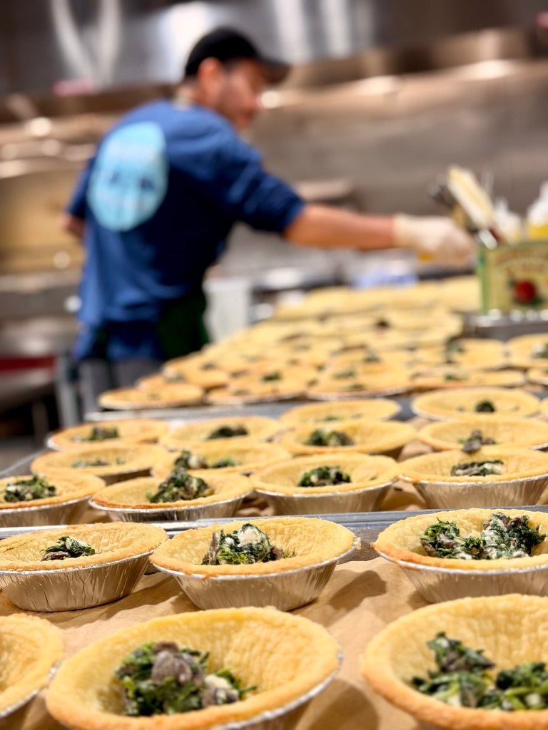 Trays of spinach and mushroom tarts lined up on the bench while a cook works behind them