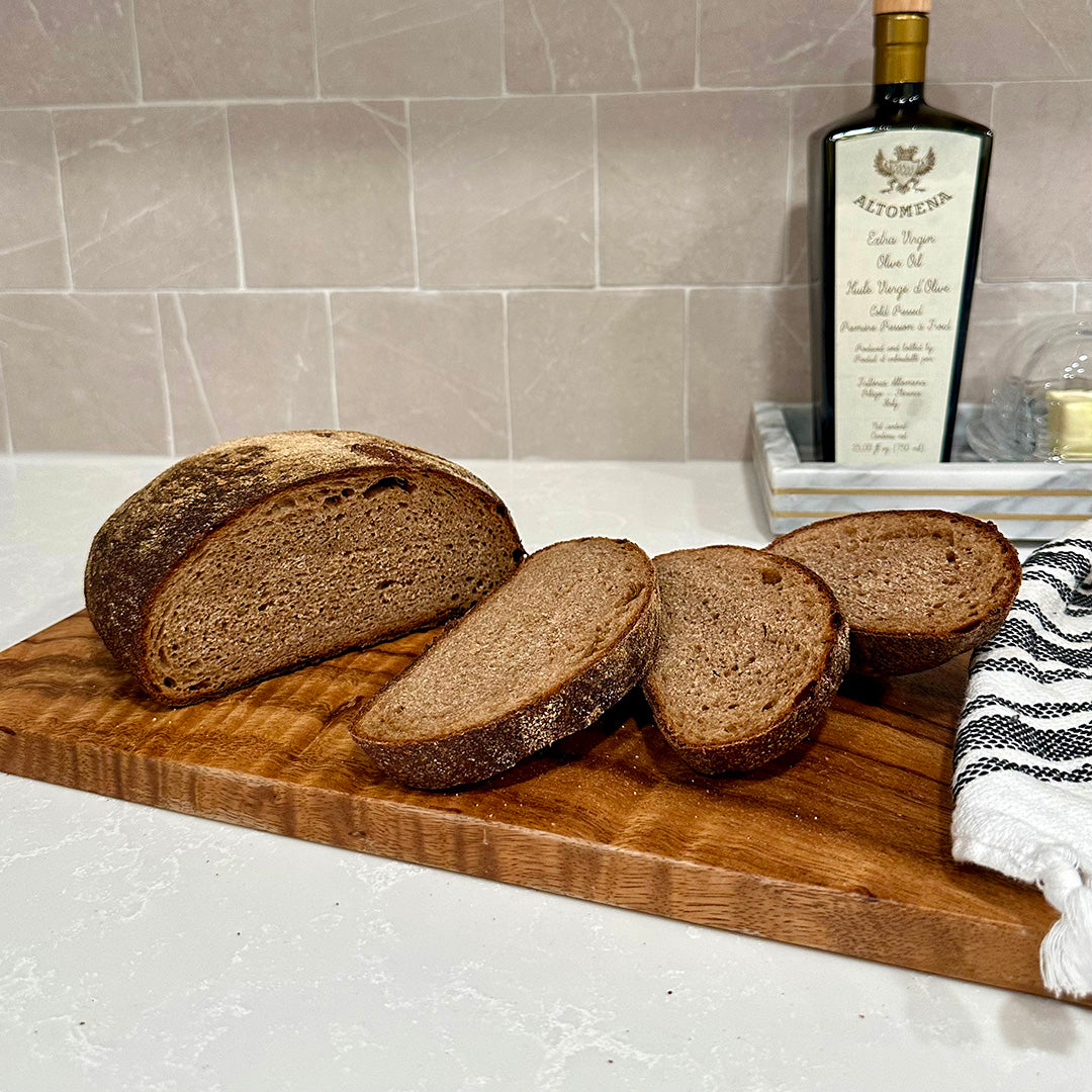 Sliced whole wheat bread on a wooden cutting board with a loaf partially sliced in the background, with a bottle of olive oil and a kitchen towel in the setting.