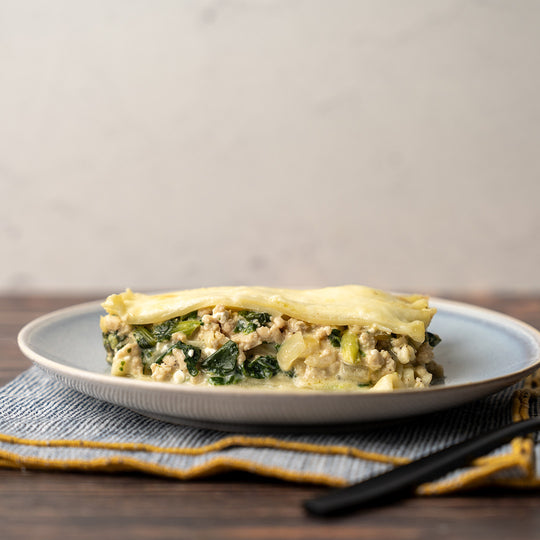 A plate containing a slice of white chicken lasagna with spinach on a wooden table with a blue and yellow napkin.