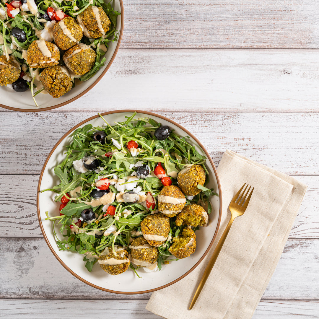 Two plates of falafel salad with tahini dressing, consisting of green falafel balls on top of a salad greens base, garnished with chopped vegetables, feta cheese, and olives, served with a side of pita bread.