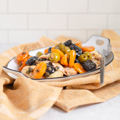 A plate of Chicken Marbella with boneless chicken thighs, apricots, plums, capers, and green olives on a yellow cloth.