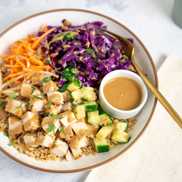 A prepared Thai Chicken Bowl consisting of roasted chicken breast, brown rice, and vegetables including purple cabbage, carrots, bean sprouts, and edamame, served with a side of peanut sauce.