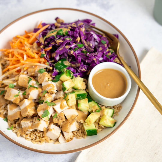 A prepared Thai Chicken Bowl consisting of roasted chicken breast, brown rice, and vegetables including purple cabbage, carrots, bean sprouts, and edamame, served with a side of peanut sauce.