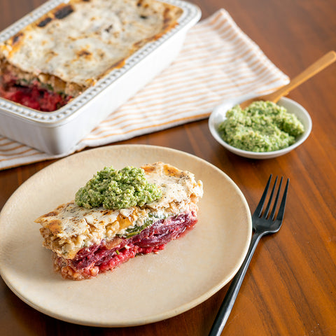 A plate with a slice of roasted beet lasagna topped with a green pesto-like sauce, with the余ing lasagna in the background.