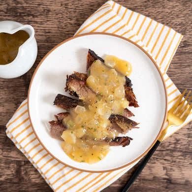 A plate of beef brisket with balsamic glazed cipollini onions on a wooden table with a yellow and white striped napkin.
