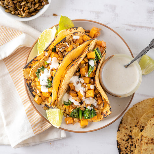 A plate with three tacos filled with roasted yam, kale, and onions, topped with a white sauce and lime on the side.