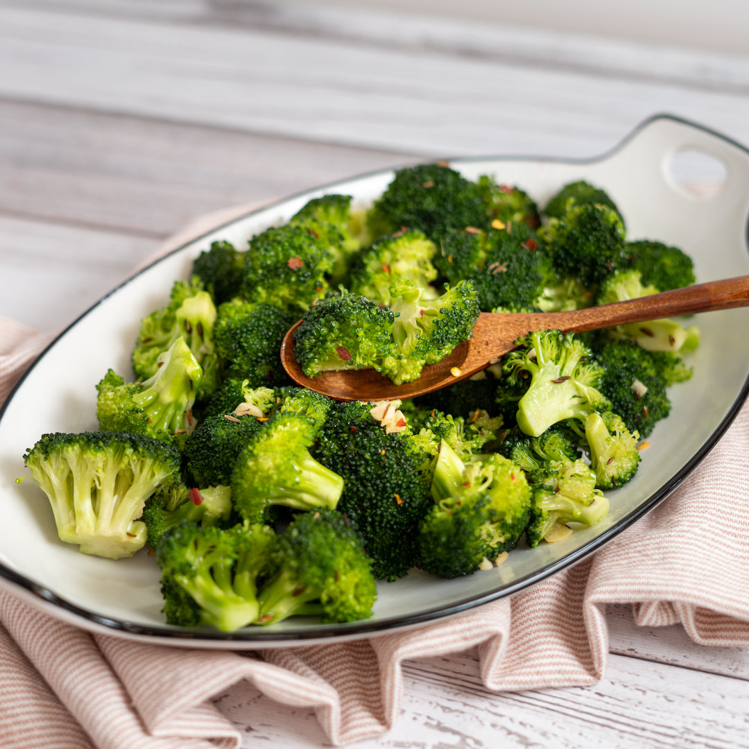 Broccoli florets served on a white oval platter with a black rim, with a wooden spoon lifting some of the broccoli. 