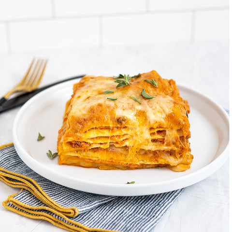 A serving of lasagna on a white plate, garnished with herbs, with a fork and napkin beside it.