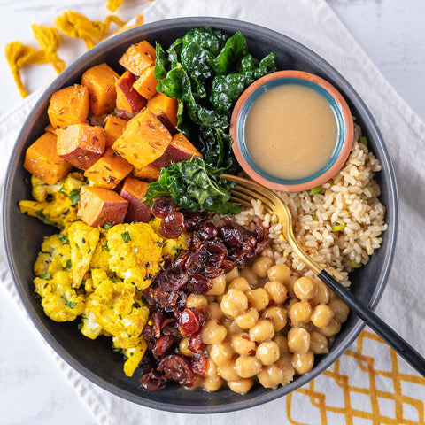 A prepared grain and roasted veggie bowl with chickpeas, sesame brown rice, roasted yams, cauliflower, orange cranberries, and kale, drizzled with tahini vinaigrette.