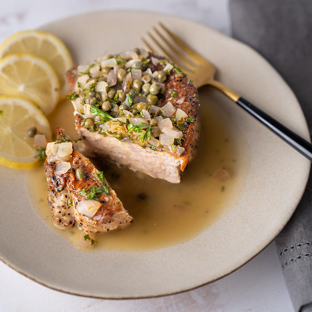 A plate with a pork chop topped with a lemon caper sauce, alongside lemon slices and a fork.