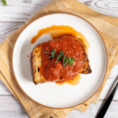 A piece of meatloaf with marinara sauce on top, garnished with a sprig of thyme, served on a white plate with a beige napkin underneath.