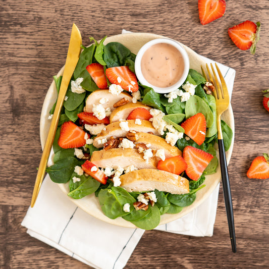 A plated salad consisting of roasted chicken, fresh strawberries, pecans, and gorgonzola cheese on a bed of spinach, served with a side of strawberry poppyseed dressing.