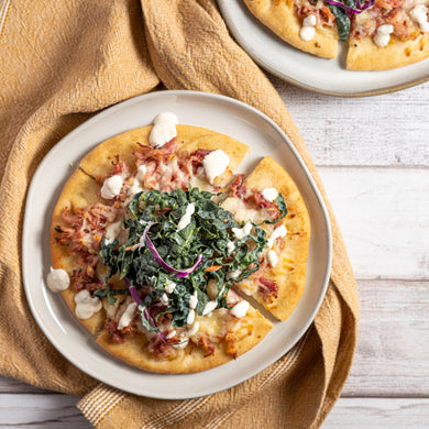 A plate of grilled cider braised pork flatbreads with kale slaw and horseradish aioli on top, served on a white plate with a yellow cloth underneath.