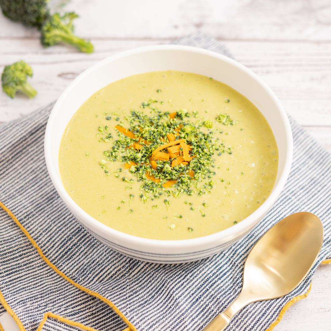 A bowl of broccoli cheddar soup garnished with chopped broccoli and cheese, placed on a striped napkin with a spoon beside it.