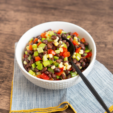 A bowl of black bean and corn salad with red bell pepper, dressed with a zippy dressing, served on a wooden table with a striped napkin.