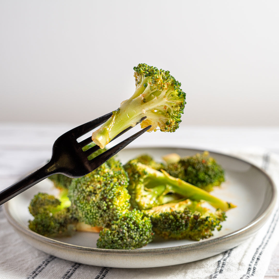 A plate of roasted broccoli florets drizzled with sriracha sauce, on a white plate with a fork holding a piece of broccoli.