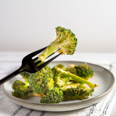 A plate of roasted broccoli florets drizzled with sriracha sauce, on a white plate with a fork holding a piece of broccoli.