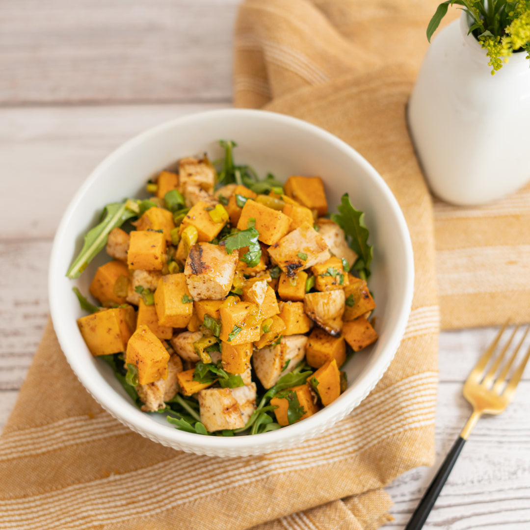 A bowl of grilled chicken and sweet potato salad with chipotle vinaigrette on a wooden table with a striped napkin, a fork, and a small vase in the background.