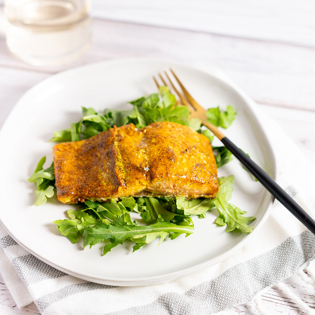 A piece of salmon seasoned with Moroccan Ras el Hanout spice blend, served on a bed of leafy greens on a white plate with a fork and knife.