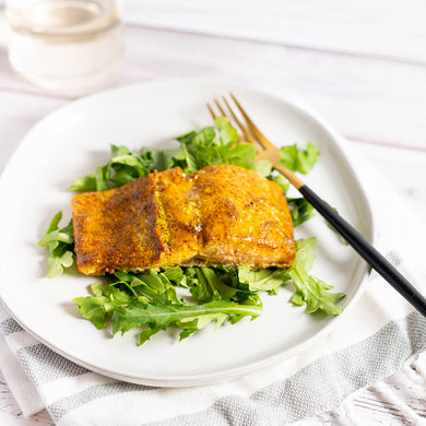 A piece of salmon seasoned with Moroccan Ras el Hanout spice blend, served on a bed of leafy greens on a white plate with a fork and knife.