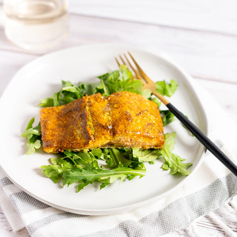 A piece of salmon seasoned with Moroccan Ras el Hanout spice blend, served on a bed of leafy greens on a white plate with a fork and knife.