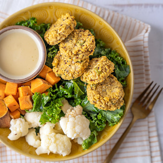 A protein-packed veggie bowl with sautéed kale, curried lentil and quinoa patties, cumin-roasted yams, and pickled cauliflower, served with a side of creamy dressing.