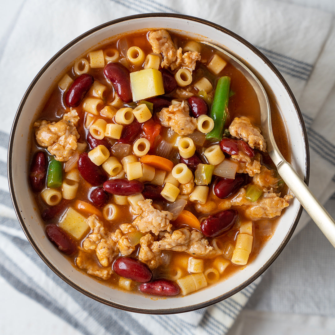 A bowl of minestrone soup with sausage, beans, and pasta, with a spoon on the side.